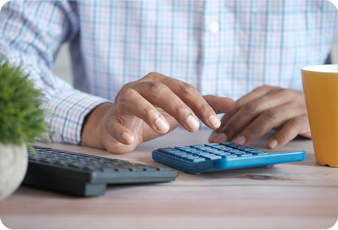 Person in checkered shirt using blue calculator at office desk with laptop, coffee cup, and small plant for financial planning and budgeting